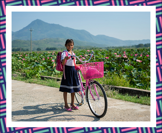 A young girl walking a bike in front of a green field and mountain range.