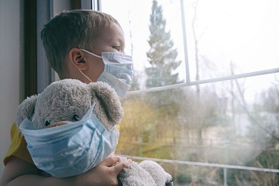young boy wearing mask holding teddy bear looking out window