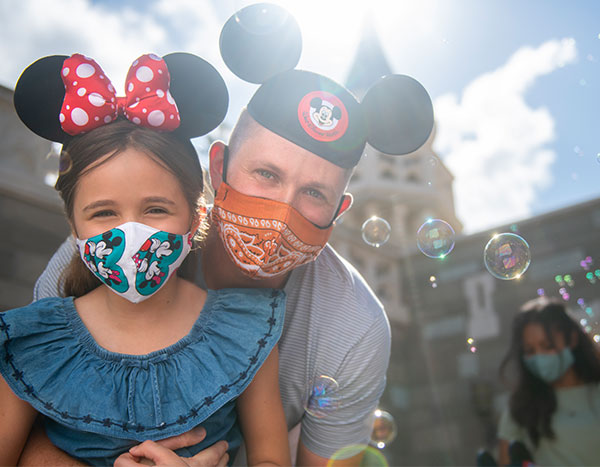 A father donning a Mickey ears hat and a face mask embraces his young daughter who is wearing Minnie ears and a Mickey & Minnie face mask.