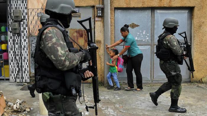 TOPSHOT - A mother and child walk past military police on patrol near the Vila Kennedy favela in Rio de Janeiro on February 23 2018 More than 3 000 soldiers supported Rio de Janeiro police Friday in a sweep of three violence-plagued favelas in the west of the Brazilian city officials said Brazilian President Michel Temer handed the military full control of security in Rio de Janeiro in an increasingly desperate fight to tame runaway gang violence AFP PHOTO CARL DE SOUZA