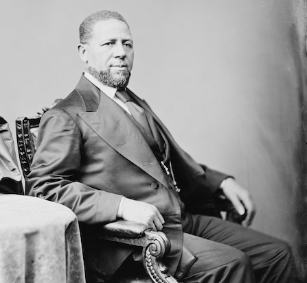 Hiram Rhodes Revels wears a suit and is seated on an ornate chair in a black and white photo.