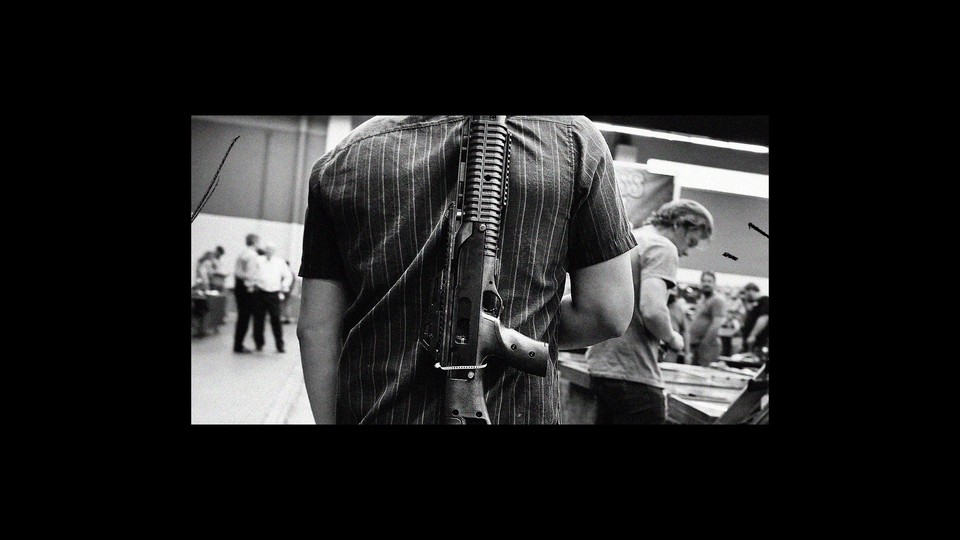 Black-and-white photo of a man carrying a rifle on his back at a gun show