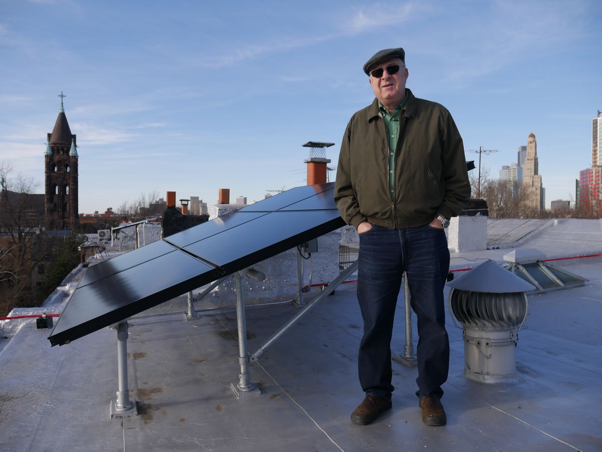 A man stands on a roof next to sloping black solar panels. The man is in outerwear. There is a city skyline in the distance. 