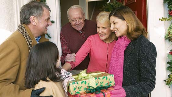 A family smiles in a doorway while a little girl holds up a stack of presents to an older woman. 