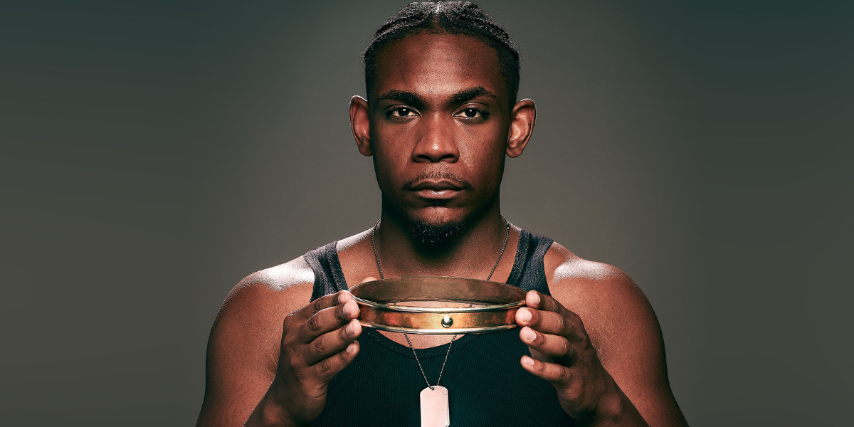 An actor with dark skin wearing a black tanktop and silver dog tag necklace faces the camera and holds a gold crown in his hands