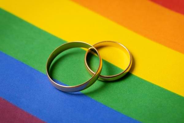 Two overlapping wedding rings sit on top of an LGBTQ pride rainbow background. © By ronstik/stock.adobe.com