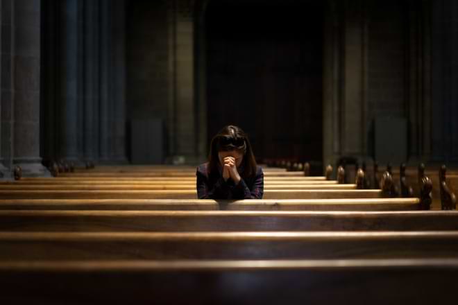 Alone at church, a woman sits in a pew, her head bowed and her hands folded in prayer. © By No-Te/stock.adobe.com