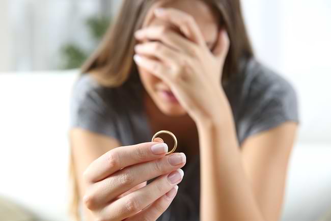 A distraught woman, her hand over her face, holds her wedding ring out in front of her. © By Antonioguillem/stock.adobe.com