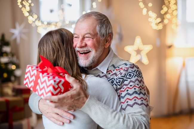 A grandpa holds a Christmas present in one hand as he hugs a woman. © By Halfpoint/stock.adobe.com