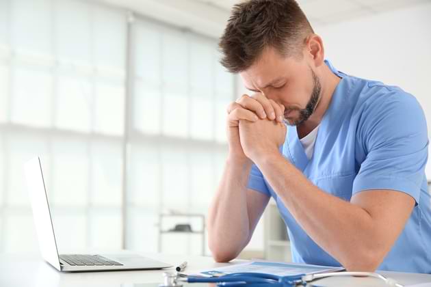 A doctor in blue scrubs sits at a desk in front of an open laptop with his hands clasped in prayer. © By Pixel-Shot/stock.adobe.com