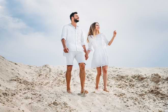A man and woman, both dressed in white summer clothes, hold hands as they walk barefoot on a sandy dune. By LIGHTFIELD STUDIOS/stock.adobe.com