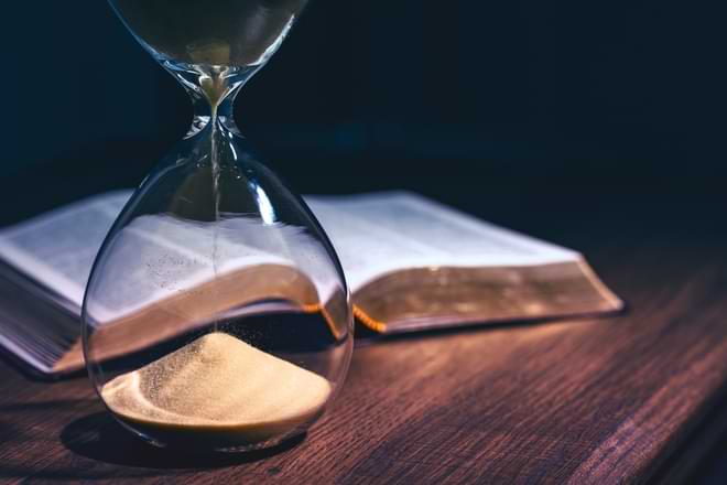 An hourglass that's almost out of time stands in front of an open Bible on a desk. By JavierArtPhotography/stock.adobe.com