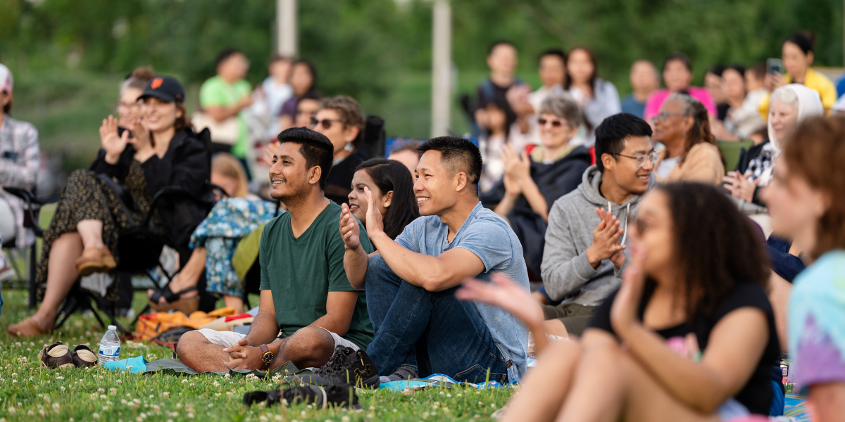Crowd of people sitting in the grass at an event, applauding and smiling
