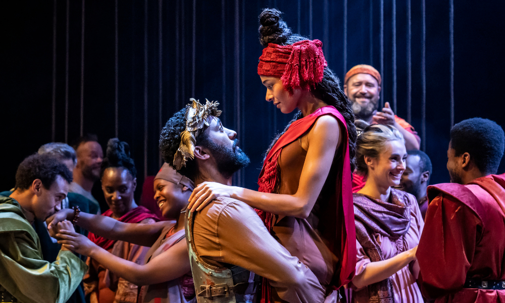 An ensemble of actors dressed in Greek clothing, with man in center holding up a woman in his arms