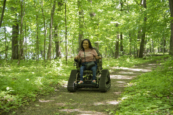 A woman in an all-terrain track chair navigates the chair through a green forest
