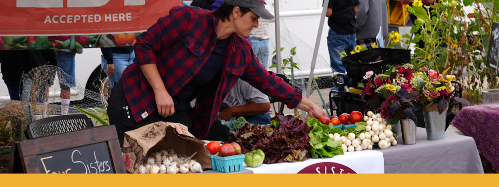 person in red and navy flannel and dad cap re-arranging tomato display at their booth. Onions, lettuce, and flowers are also visible on the table.