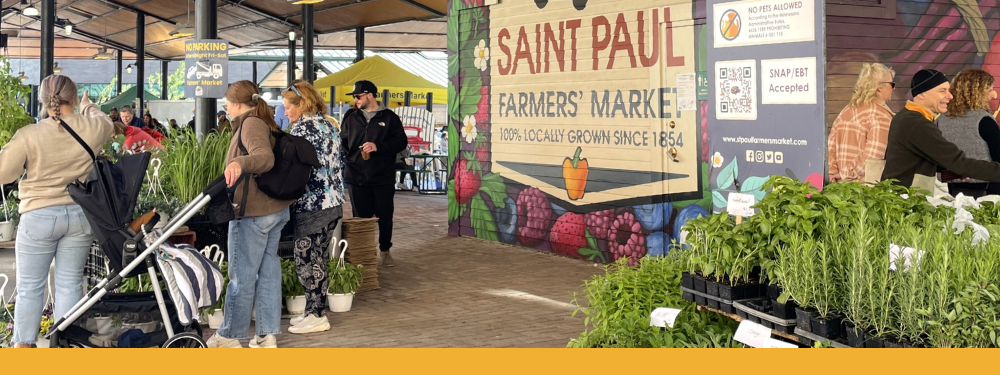 a crowd admiring a vegetable stand next to a large painted wall reading, "SAINT PAUL FARMERS' MARKET"