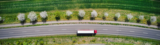 overhead photo of single red and white lorry truck driving on a road surrounded by green