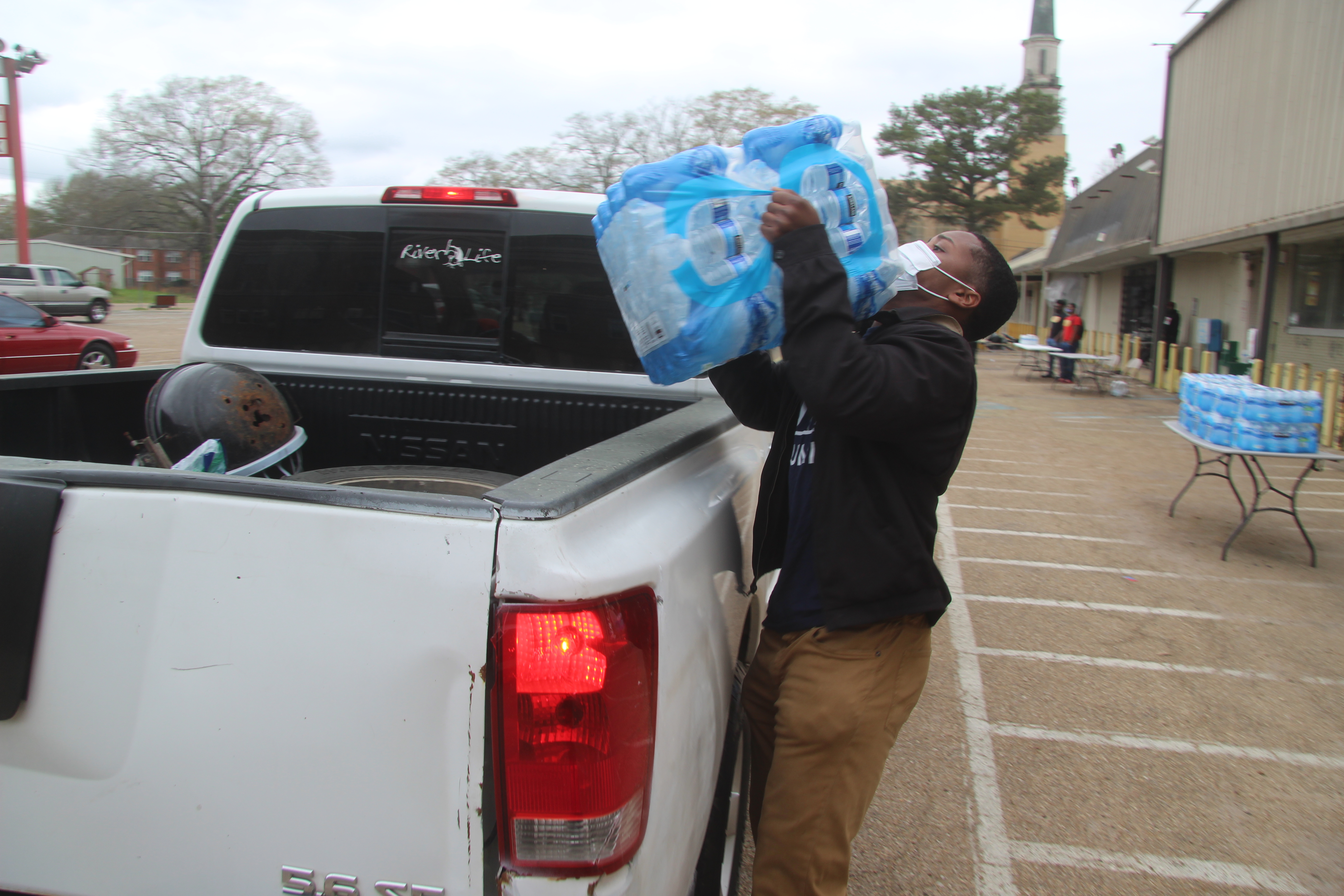 A volunteer wearing a powder blue face mask, black jacket, and brown pants hoists a case of water into the back of a white pick -up truck. In the background is a table with additional cases of water.
