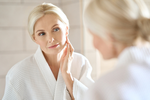 Headshot of gorgeous mid age adult 50 years old blonde woman standing in bathroom after shower touching face_ looking at reflection in mirror doing morning beauty routine. Older skin care concept.
