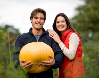 Young smiling couple standing on allotment holding large pumpkin smiling at camera