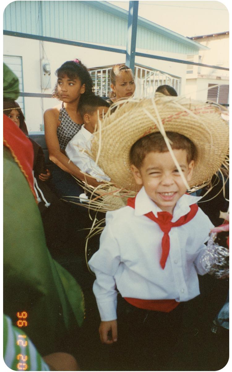 A grainy photograph of a young boy in a straw hat and Puerto Rican jibaro outfit gives a mischievous smile and wink to the camera