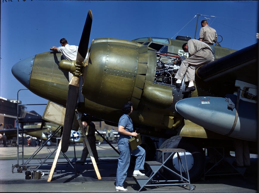 Color photograph of a group of people working on an airplane. Several people are on the top and sides of the aircraft; a woman is starting to climb the stairs next to the aircraft.