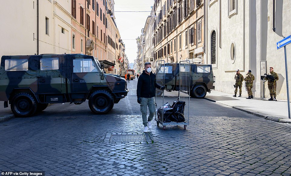 A man in a face mask pulls a cart along a street in Rome under the watchful eyes of Italian soldiers on the pavement today