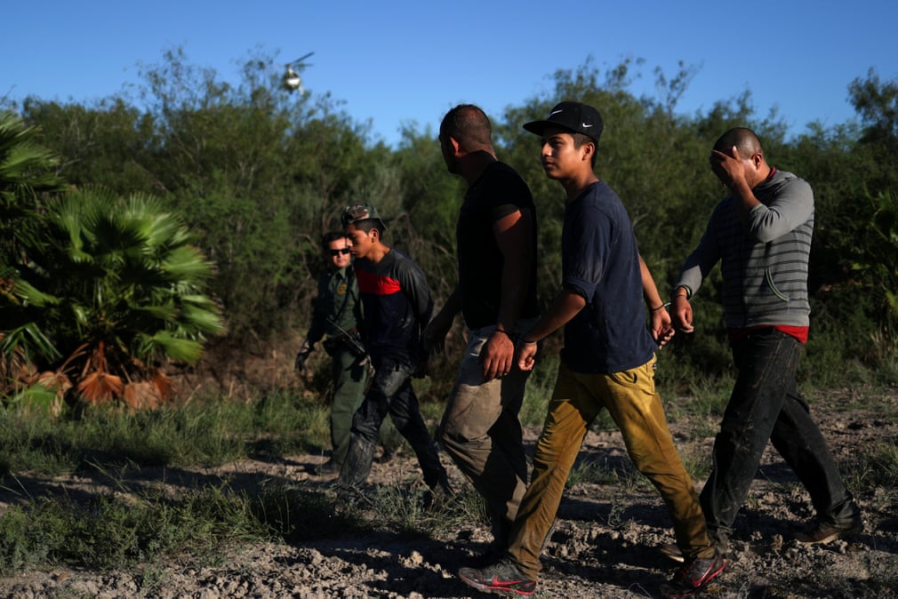 A helicopter with the U.S. Customs and Border Protection’s (CBP) Air and Marine Operations (AMO) searches in the background as a group of men from Central America are apprehended for illegally crossing into the United States from Mexico in Penitas, Texas
