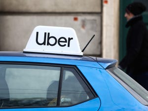 An 'Uber' placard sits on top of a car roof and a person walks past in the background.