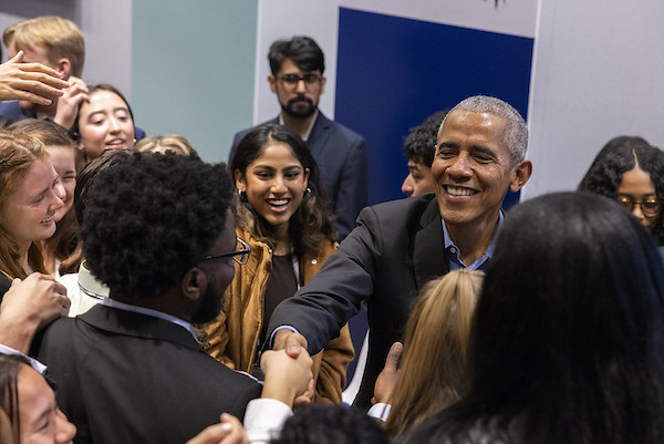 President Obama is smiling and surrounded by young, smiling people with light to dark skin tones. He is shaking hands with one of the people near him.