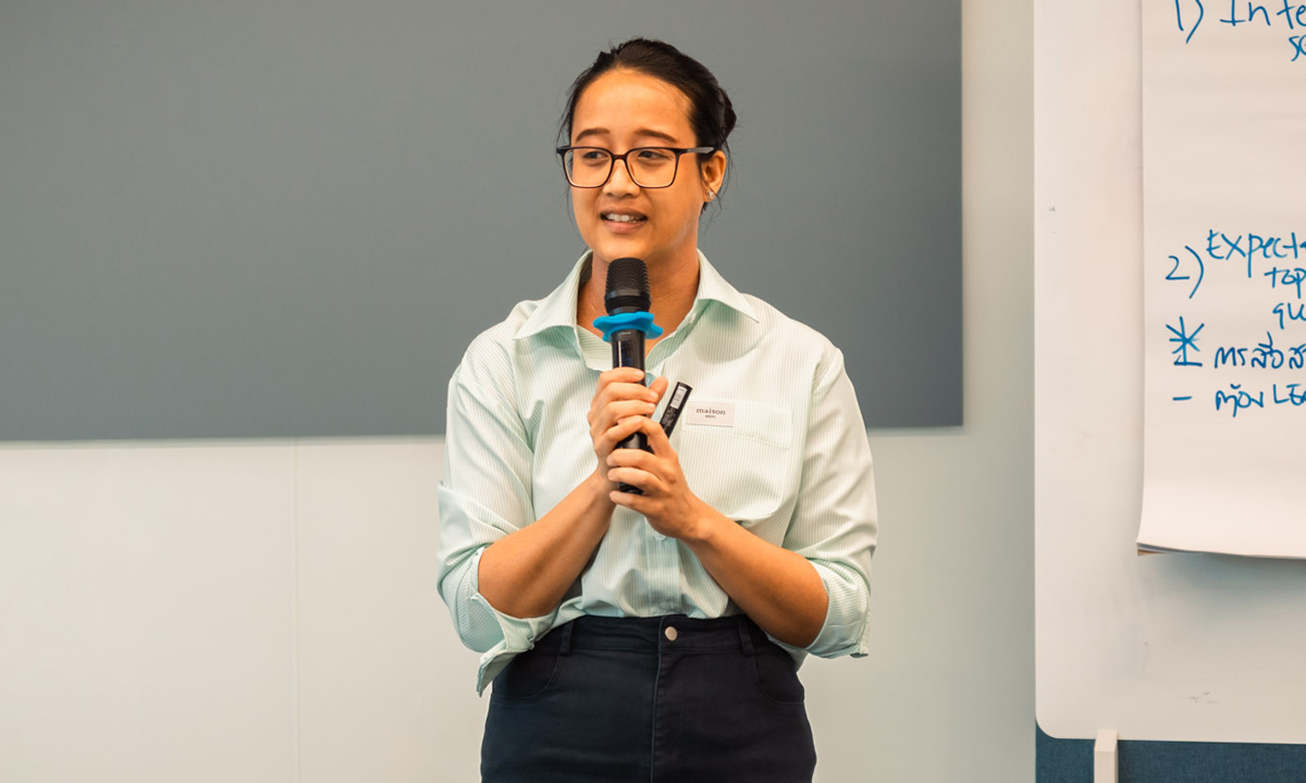 A woman with an olive skin tone and glasses holds a microphone and stands speaking at the front of a room.