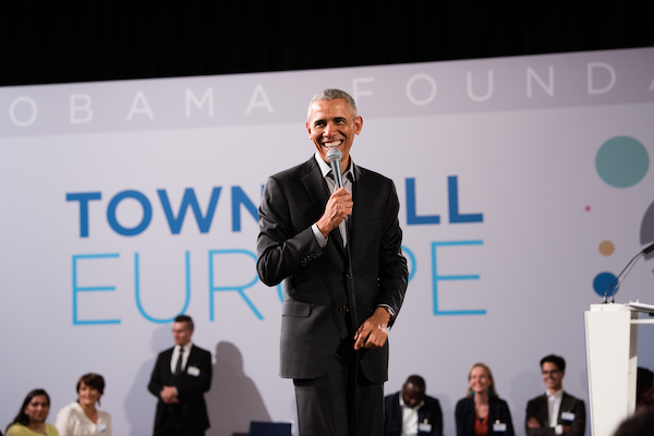 President Obama is smiling and holding a microphone while addressing attendees for a town hall in Europe. There are several people with light to dark skin tones behind him listening.