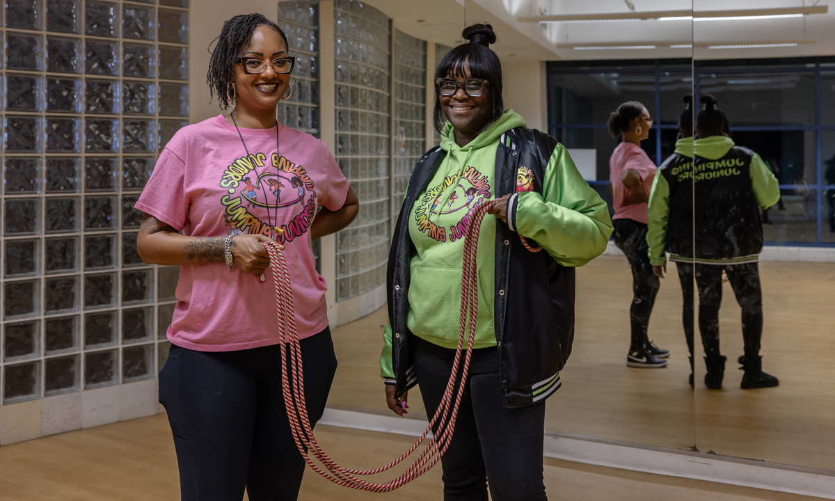 Two women, with a medium skin tone and a medium-dark skin tone, stand smiling with a jump rope held between them