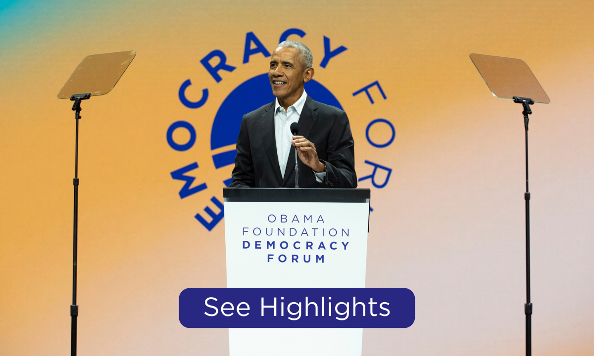 President Obama stands behind a podium as he speaks at the Obama Foundation Democracy Forum.  The president has a medium skin tone and is wearing a black suit. In the background is a blue, green, and yellow gradient graphic that reads, “Democracy Forum.” At the bottom of the graphic is a blue rectangular button that reads, “see highlights.” 