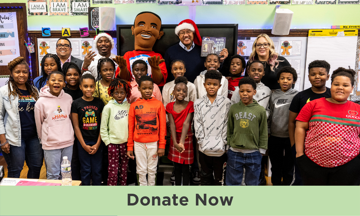 A group of young children of varying skin tones pose for class photo in their classroom with Mike Strautmanis in the back, who is wearing a Santa hat. A mascot of a man with a deep skin tone wears a red shirt and waves to camera. “Donate Now” is written in black text against a mint border at the bottom of the image.