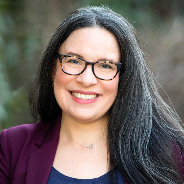 Angela Barranco, a woman with a light medium skin tone, smiles at the camera. She is standing outside and is wearing glasses and coral lipstick. She has long salt and pepper hair.