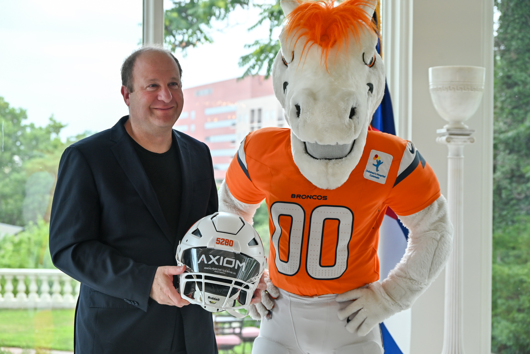 Governor Polis stands next to the Broncos mascot, holding a Broncos football helmet.
