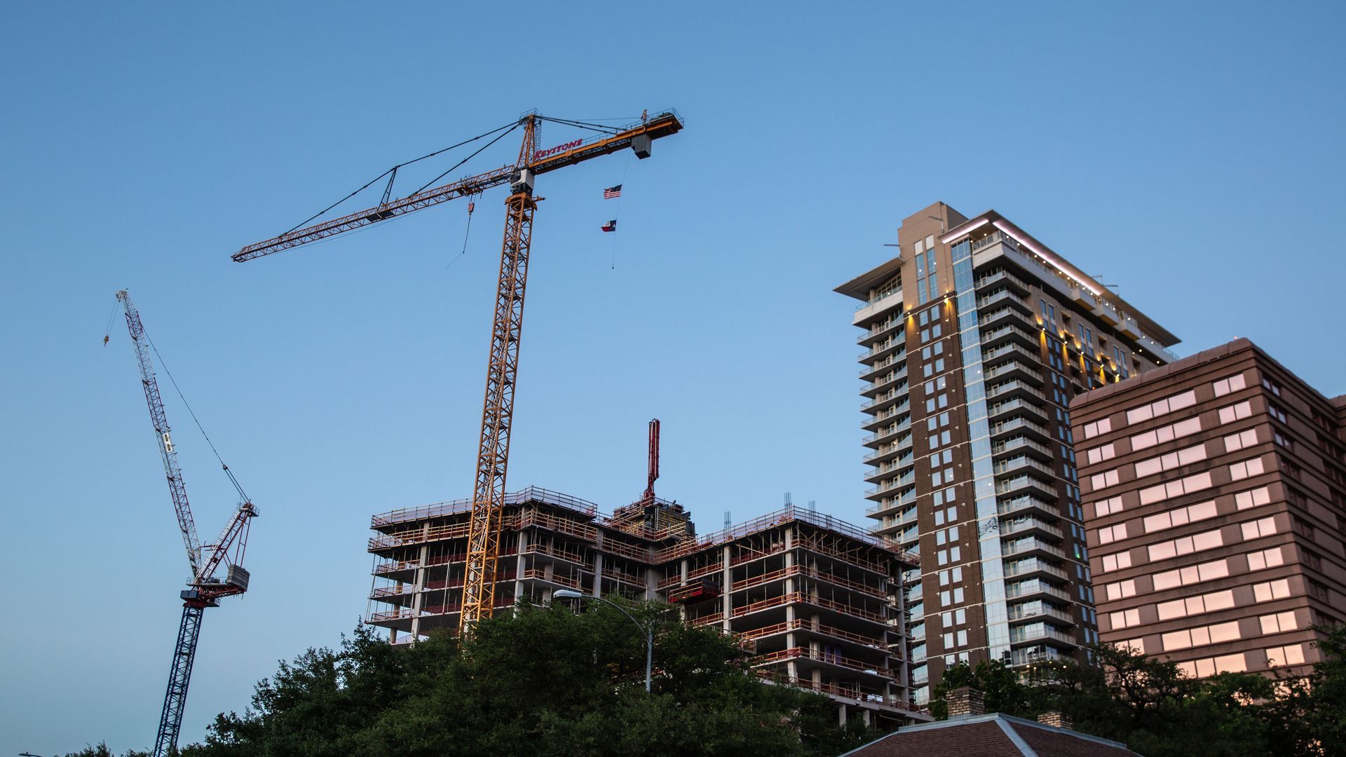 A photo showing two cranes in front of an apartment building that’s under construction next to a taller apartment building that is complete. The sky looks clear and tops of trees are visible in the foreground. 