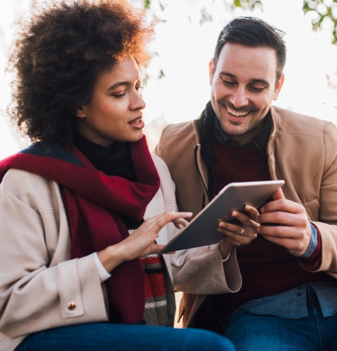 A couple sitting on a park bench, with a tablet in hand, happily discussing factors that could affect their mortgage rate.