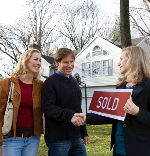 A couple standing outside, with their new home in the background, being congratulated by their real estate agent.