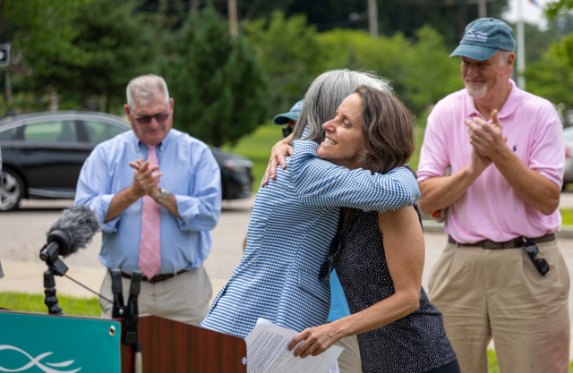 Screenshot_(60).png Democratic Whip Clark hugs Executive Director of the Charles River Watershed Association Emily Norton in front of a podium. Behind them, state and local officials applaud.