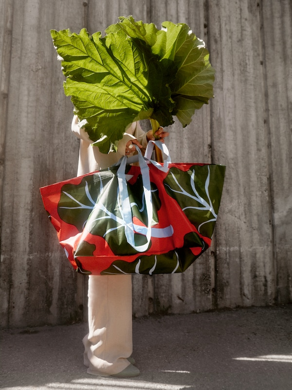 A person is holding a BASTUA carrier bag with a leaf pattern and two large rhubarb leaves to hide their face.