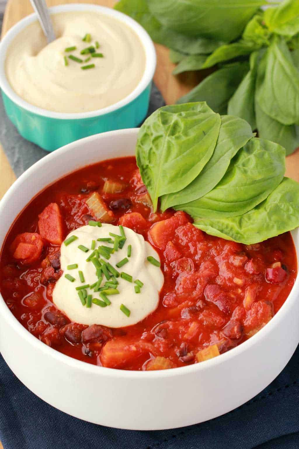 Vegan Chili in a white bowl with a dollop of cashew cream, chopped chives and fresh basil leaves. A bowl of cashew cream and more fresh basil is in the background. 
