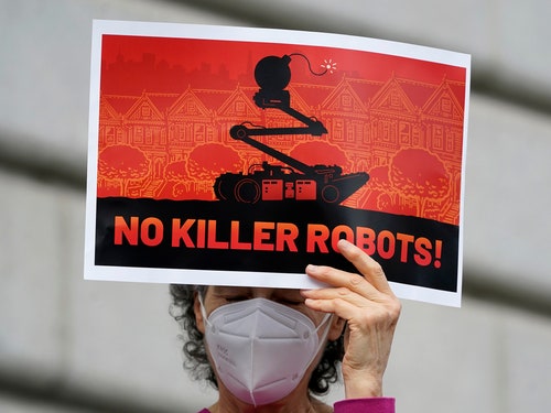 Person wearing a face mask holds a sign that says "No Killer Robots!" with an illustration of a robot with a bomb on top.