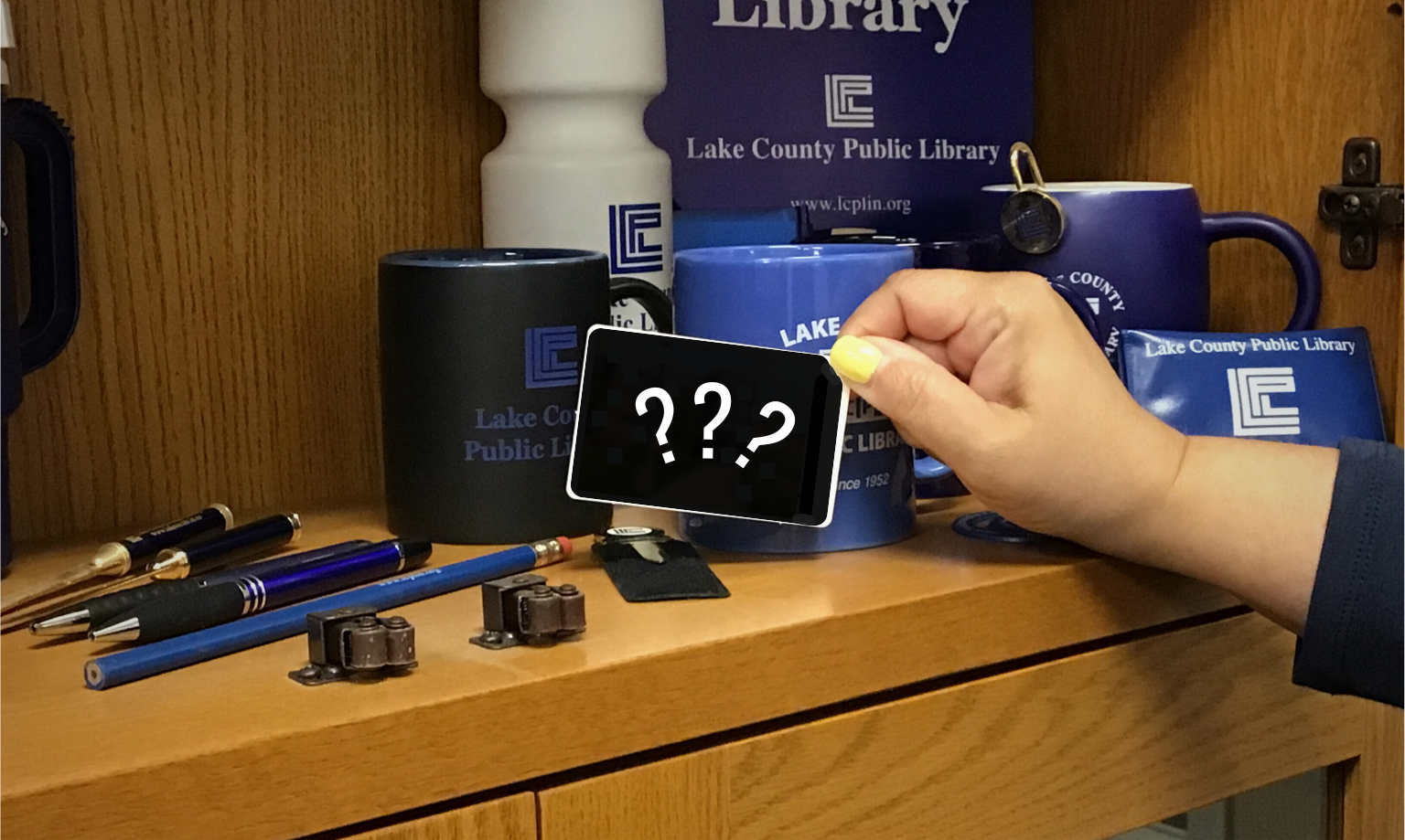 A hand holds up a library card, but the design on it has been blotted out and replaced with question marks. It's being held in front of a shelf of old library swag featuring different logos and slogans