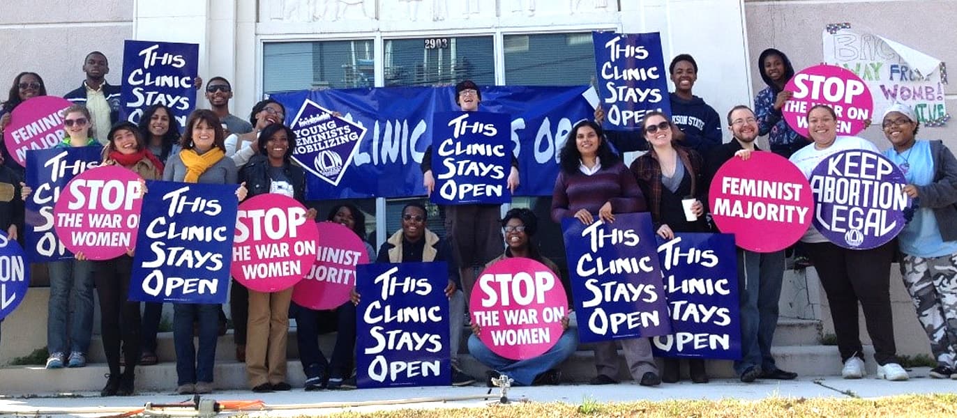 Photo of the National Clinic Access Project team, a group of 21 people holding signs that read 'This Clinic Stays Open', 'Keep Abortion Legal', 'Stop the War on Women', and 'Feminist Majority'