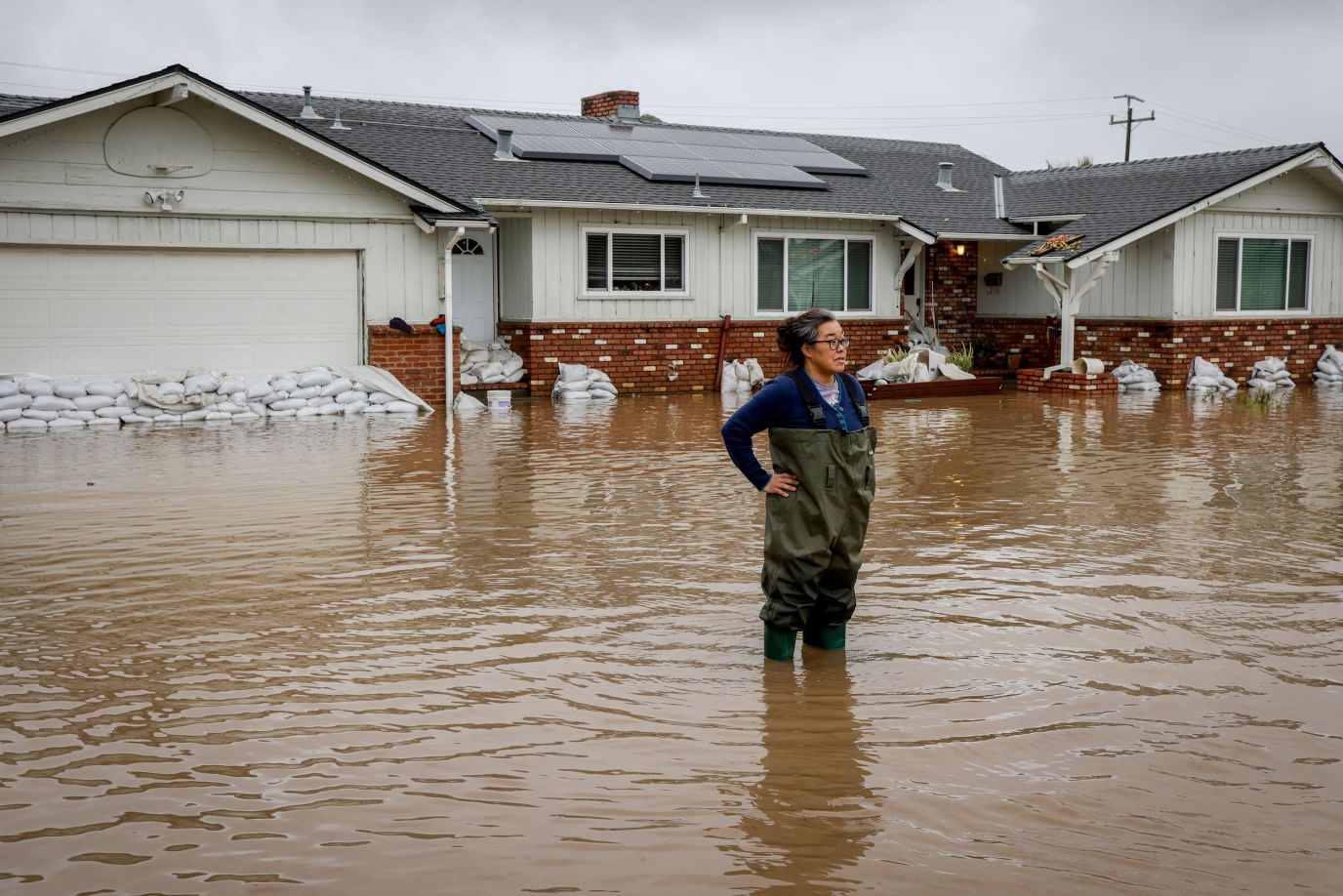A Californian stands knee deep in brown floodwater in front of a solar-powered home with sandbags