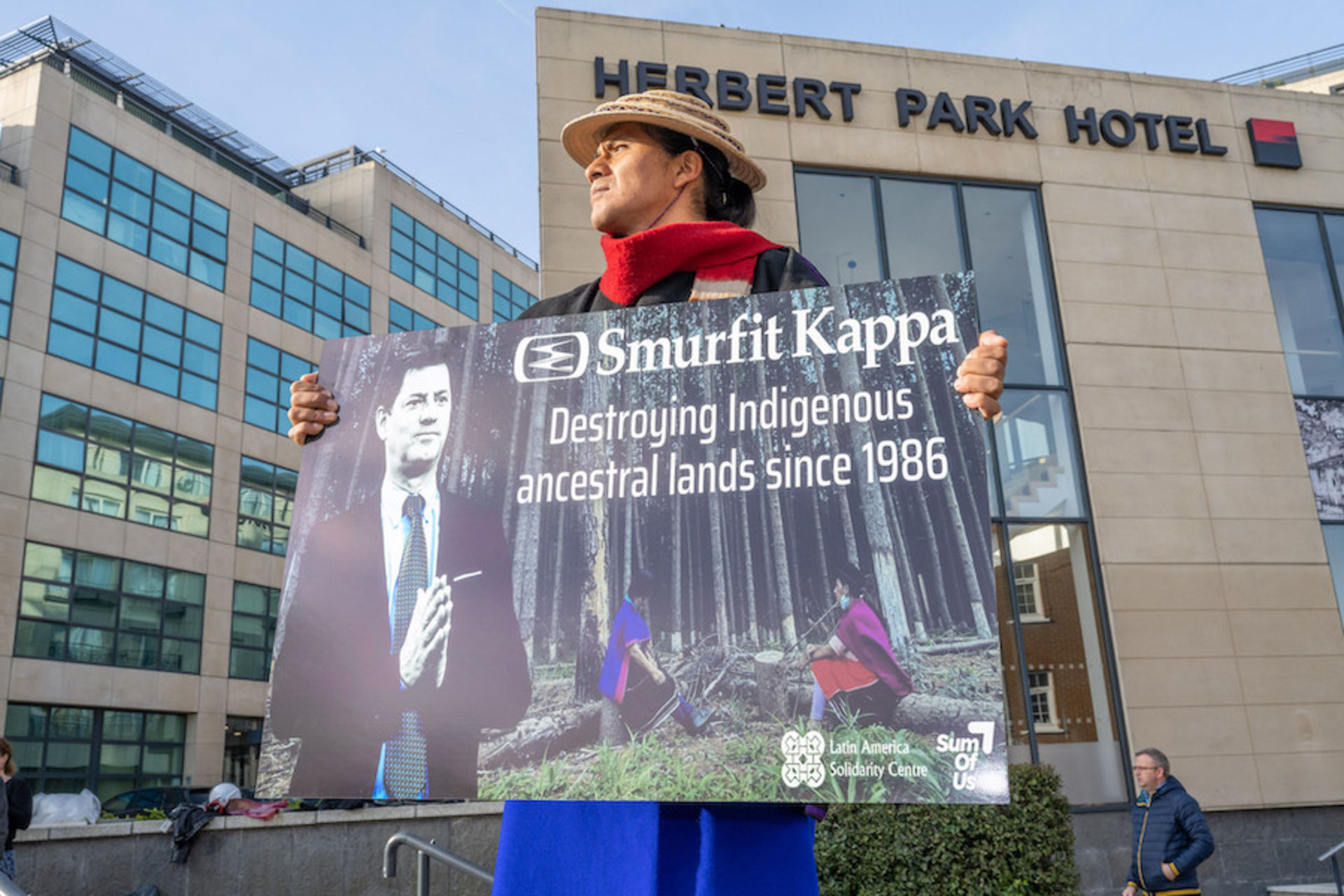 Pedro, chapeau sur la tête et écharpe rouge au cou, tient une affiche dénonçant la destruction causée par Smurfit Kappa devant le lieu dans lequel a eu lieu l'assemblée générale de l'entreprise à Dublin.