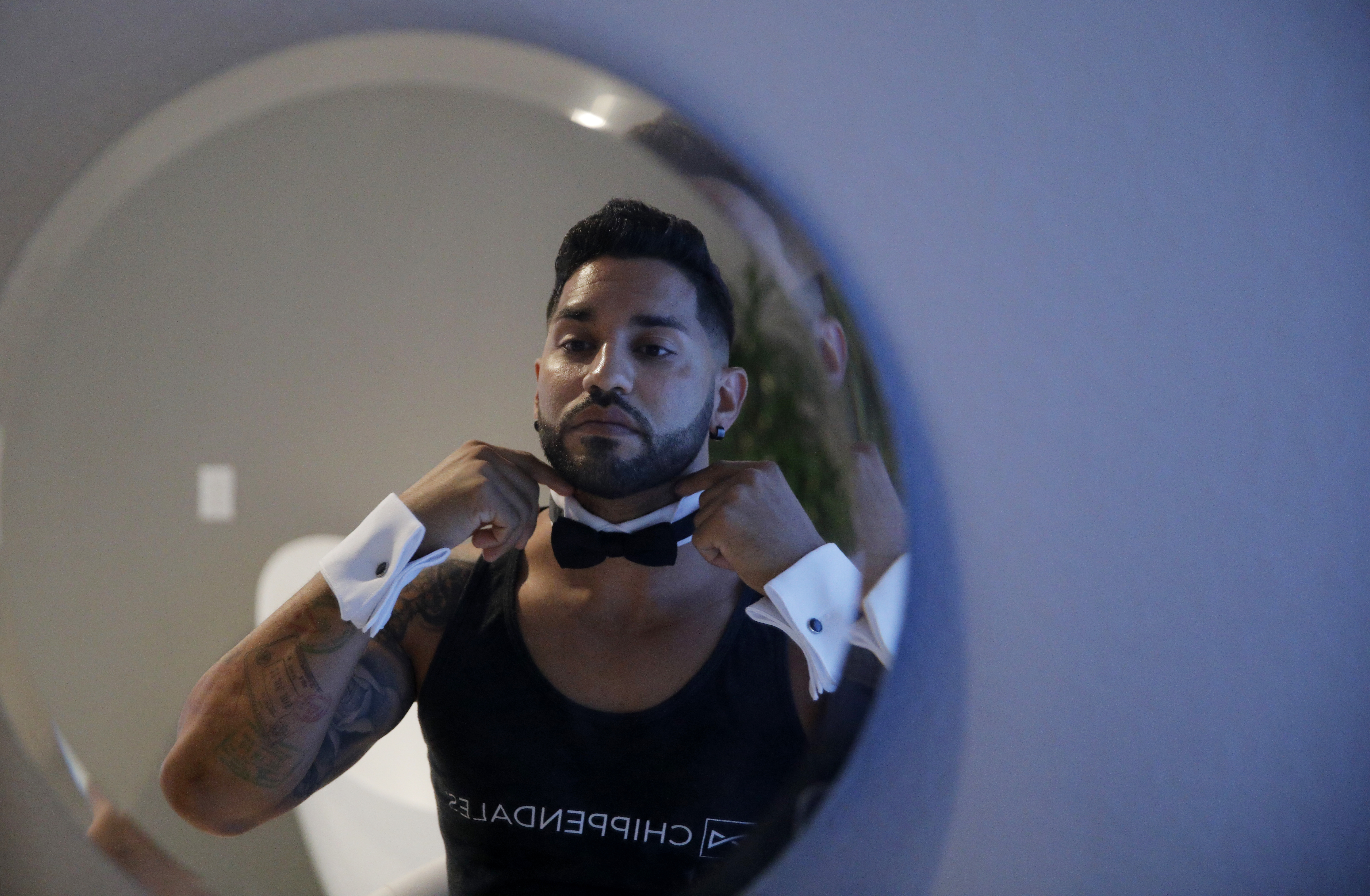 A Chippendales dancer wearing dress shirt cuffs adjusts a tuxedo collar in the mirror.